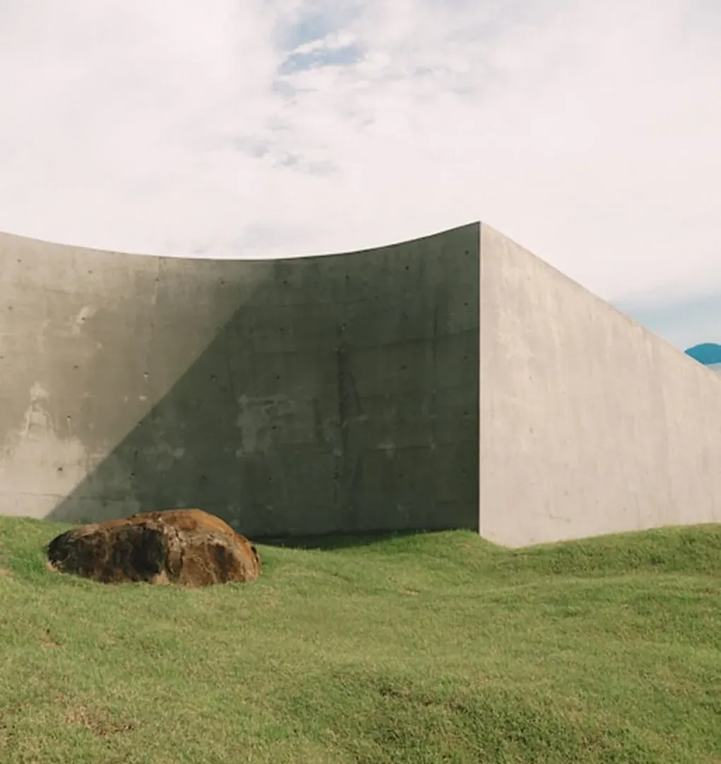 Muro curvo in cemento su prato verde con masso in primo piano e cielo chiaro